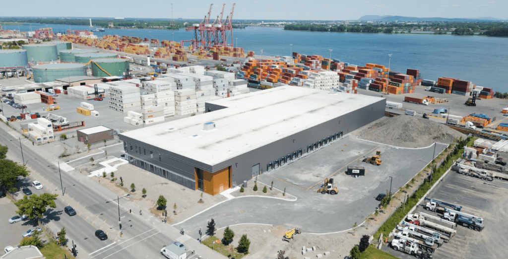 An ariel image of a newly constructed industrial building a 6100 Rue Notre-Dame Est. Montreal, Quebec, with an orange enterance, along the port and shipping yard. 